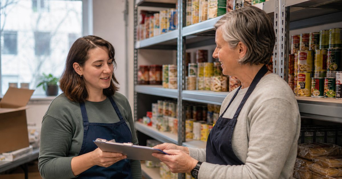 Two adult volunteers in aprons standing beside a food pantry shelving unit, one handing a clipboard to the other, warm indoor lighting, shelves of canned goods behind them